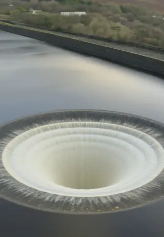 An aerial view of a circular spillway structure, resembling a giant drain, surrounded by calm water and greenery. The spillway is pulling water down into a central vortex, with a smooth edge contrasting the surrounding landscape.