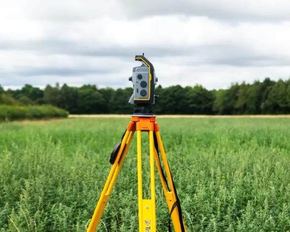 A surveying instrument mounted on a yellow tripod, positioned in a green field with trees in the background under a cloudy sky.