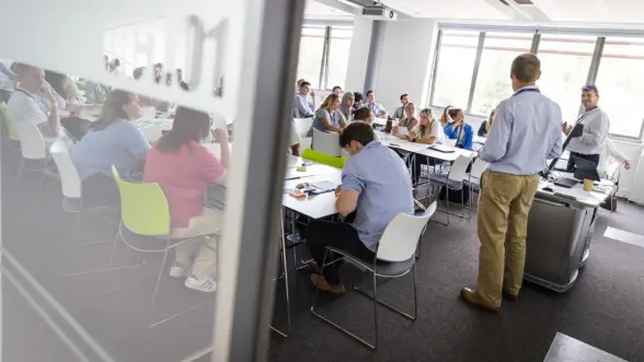 An office meeting with a speaker presenting to a large group of seated attendees. The room features modern furniture and is well-lit with large windows.