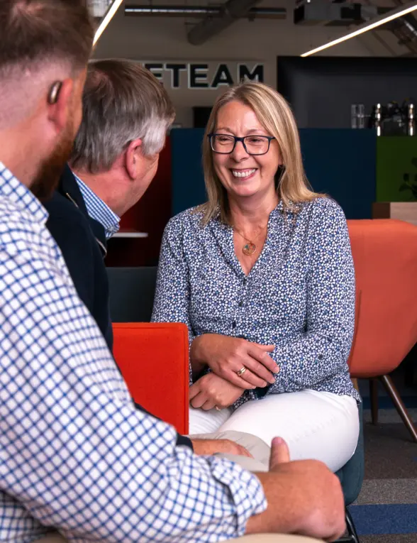 A woman with glasses smiling while sitting on a sofa, engaged in conversation with two men. One man is facing her, while the other has his back to the camera, all in a modern office setting with colorful decor.