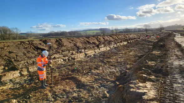 A construction worker in a high-visibility orange outfit and hard hat is using a surveying instrument on a dirt excavation site under a clear blue sky. The landscape features layered soil and distant trees.