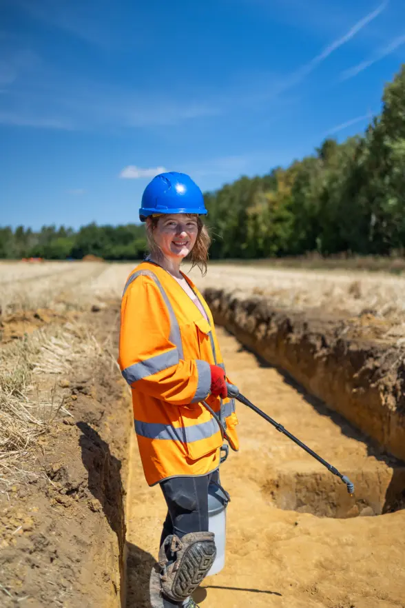 A woman in a safety helmet and orange reflective vest stands beside a trench in a field, smiling at the camera. The background shows trees and a clear blue sky.