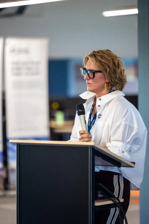 A woman with curly hair and glasses speaking into a microphone at a podium. She is wearing a white blouse and is focused on her speech, with a background that includes blurred displays.