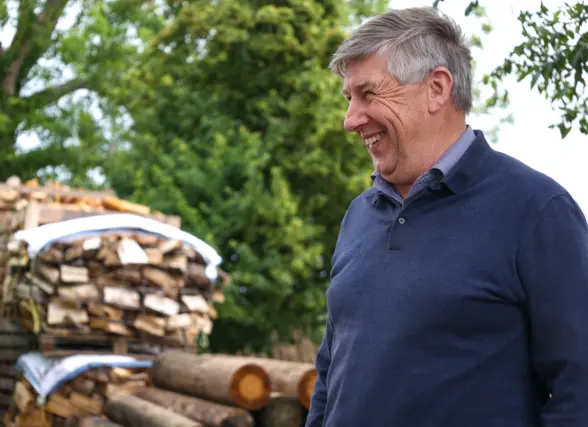 A middle-aged man smiling while standing outdoors, with a stack of firewood and logs in the background surrounded by greenery.