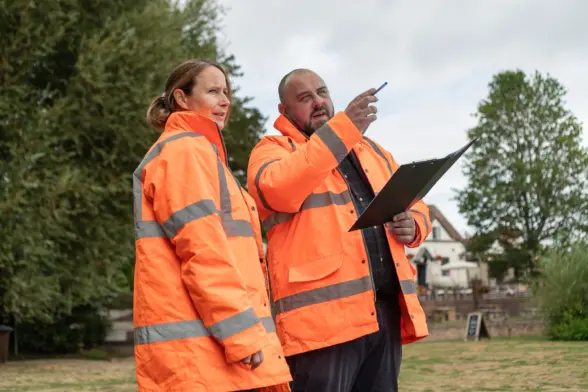 Two individuals in orange high-visibility jackets discussing a task outdoors, with one pointing while the other observes. They are standing on grass with trees and buildings in the background.