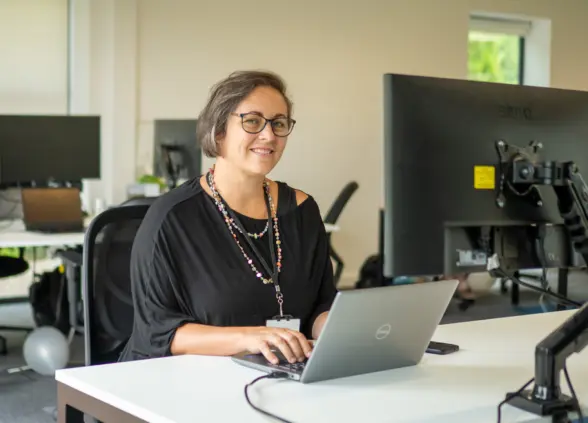 A woman with glasses seated at a desk, working on a laptop. She is wearing a black blouse and a colorful necklace, with office equipment and plants visible in the background.
