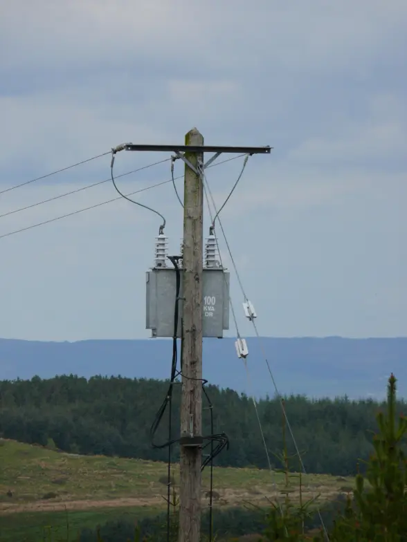 A wooden electric pole with a transformer labeled '100'. Wires are connected to the pole, and the background features a view of green hills under a cloudy sky.