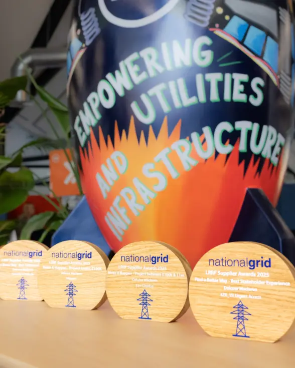 Four wooden awards from National Grid, labeled 'LGBT Supplier Awards 2023,' placed on a table in front of a colorful mural featuring the text 'Empowering Utilities and Infrastructure.'