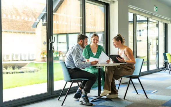 Three colleagues sitting around a table in a modern office environment, discussing documents and using a tablet. Large windows provide natural light and a view of greenery outside.
