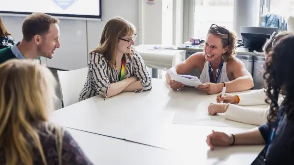 A group of six people sitting around a table engaging in a discussion. One person, who is holding papers, is speaking to the group, while others are listening attentively. The setting appears to be a modern conference room.