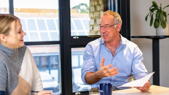 A man in a striped shirt gestures while speaking to a woman with long hair in a casual meeting setting. They are seated at a table near a window with natural light.