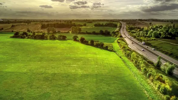 Aerial view of a lush green field adjacent to a highway, with trees lining the road. In the distance, rolling hills are under a partly cloudy sky.