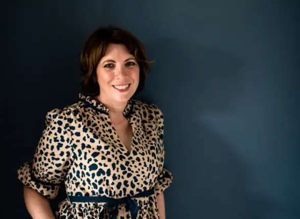 A woman standing against a dark blue wall, wearing a leopard print dress with puffy sleeves. She is smiling and has short, wavy brown hair.