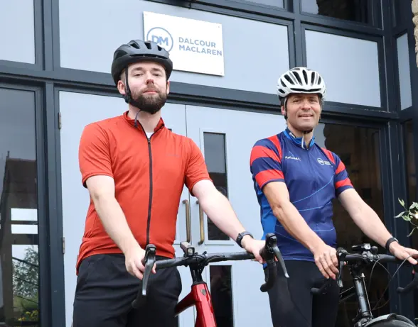 Two men in cycling gear standing with their bikes outside a building. One man wears a red shirt and black shorts, while the other wears a blue and red jersey. Both wear cycling helmets and are posing in front of a glass door with a logo.