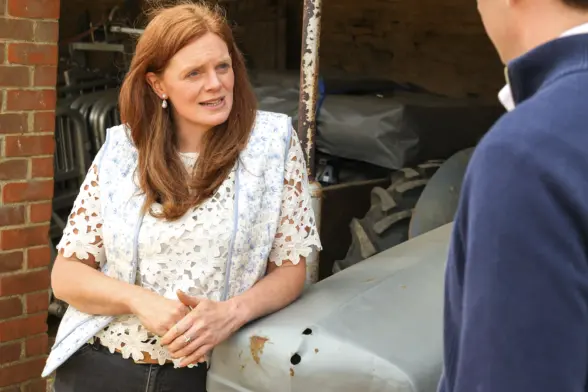 A woman with long red hair wearing a white blouse and a blue vest talks to a man in front of a tractor in a barn. The background shows various farming equipment.
