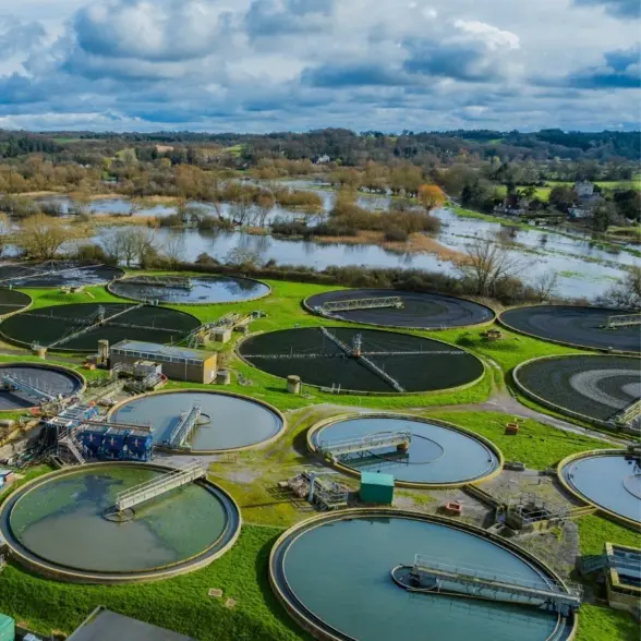 An aerial view of a wastewater treatment facility with multiple circular tanks surrounded by lush green grass. In the background, a flooded landscape with trees and a cloudy sky is visible.
