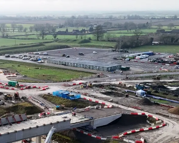 An aerial view of a construction site featuring a large parking area with several vehicles. Surrounding the site are green fields and trees. In the foreground, construction materials and red and white barriers outline the work area.