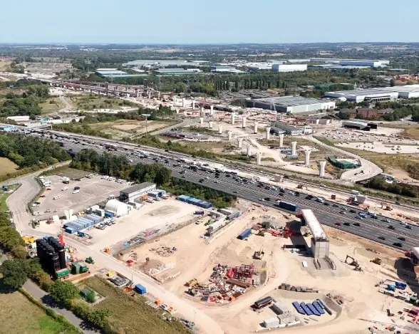 An aerial view of a construction site near a busy highway, with multiple vehicles on the road. The site features piled materials, machinery, and pillars under construction, with distant industrial buildings and green trees in the background.
