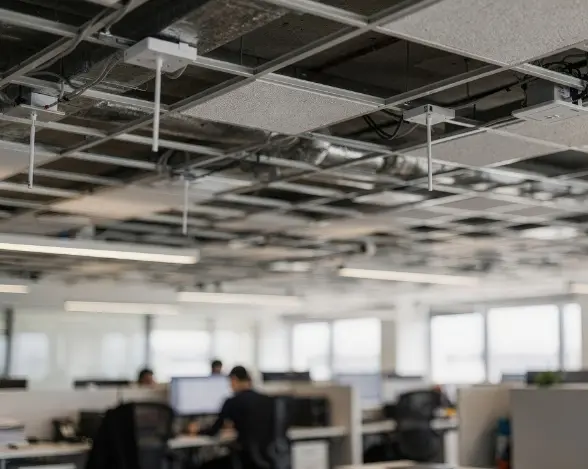 An office interior with a drop ceiling showing exposed tiles, cables, and lighting fixtures. In the background, partially visible workstations with multiple computer monitors are occupied by employees, creating a professional workspace.