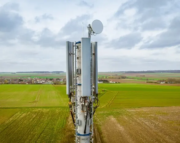 A tall telecommunications tower with multiple antennas and satellite dish stands in an open field. The surrounding landscape features green fields and patches of brown earth under a cloudy sky, with a small village visible in the distance.