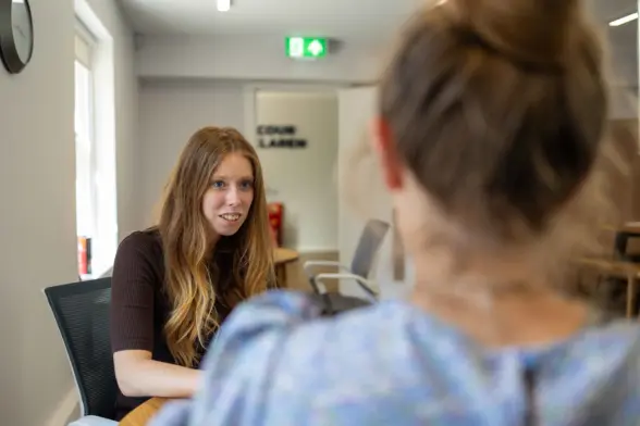Two women sit across from each other in a meeting room, with one woman in focus beside a table and the other seen from behind; a lit exit sign and doorway are in the background.