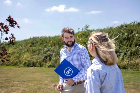 A man in a blue checkered shirt holds a blue folder marked with a white logo while talking with a woman outdoors on a grassy path beside bushes under a partly cloudy sky.