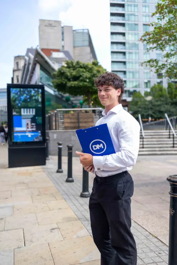 A young man in a white shirt and dark trousers holds a blue DM folder outdoors, with city buildings, steps, and a digital sign in the background.