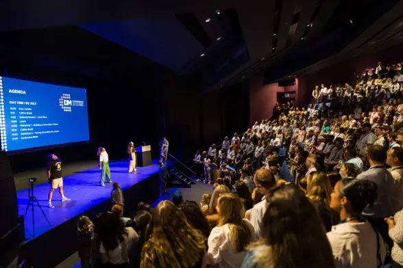 A large audience seated in a theater, facing a stage with four speakers discussing an agenda displayed on a screen. The room is well-lit, showing attentive participants.