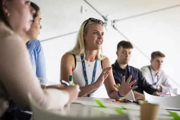 A group of people engaged in a discussion at a table, with a woman in the foreground speaking and using hand gestures. The setting appears to be a modern conference room.
