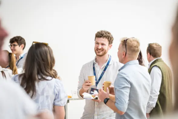 A smiling man holding two cups of coffee engages in conversation with another man at a networking event, surrounded by a small group of people in a bright, modern space.