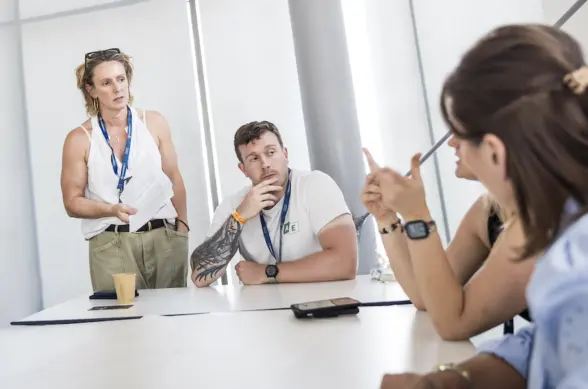 A group of professionals engaged in a discussion around a table in a bright room. One person stands and speaks while the others listen attentively, with papers and a drink placed on the table.