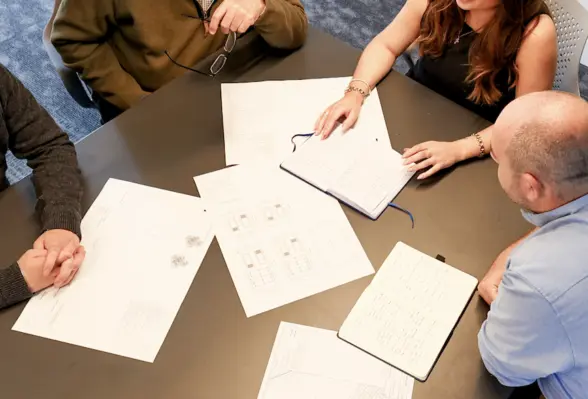 A group of professionals engaged in a discussion around a table covered with architectural plans and notebooks. Two individuals are actively taking notes while others observe the documents.