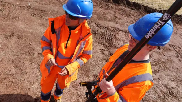 Two construction workers wearing orange high-visibility jackets and blue helmets stand on a construction site. One holds a surveying tool while discussing with the other.