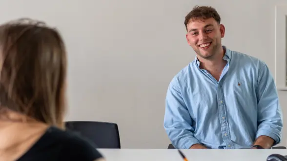 A young man in a light blue shirt is smiling while sitting at a table during a conversation. A woman with long hair is sitting opposite him, partially visible.