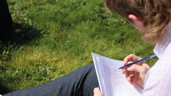 A person sitting on grass, holding a pen and writing on a notepad, with visible handwritten notes. The individual has wavy hair and wears a checkered shirt.