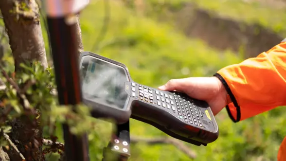 A person using a handheld data collection device in an outdoor setting, positioned near a tree with greenery in the background. The device features a touchscreen and a keyboard.
