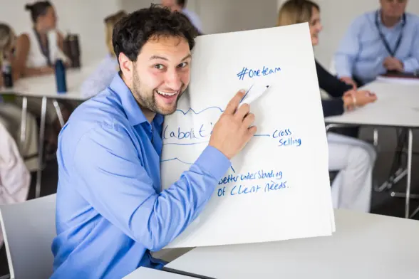 A man in a blue shirt pointing at a large paper with notes during a team discussion. The background shows colleagues seated at tables, engaged in a professional setting.