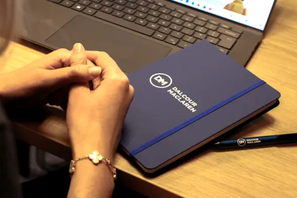A person with a silver bracelet is resting their hands on a wooden desk near a closed notebook with a blue cover and the logo "DALCOUR MACLAREN." Next to it is a black pen and a laptop with an open keyboard.