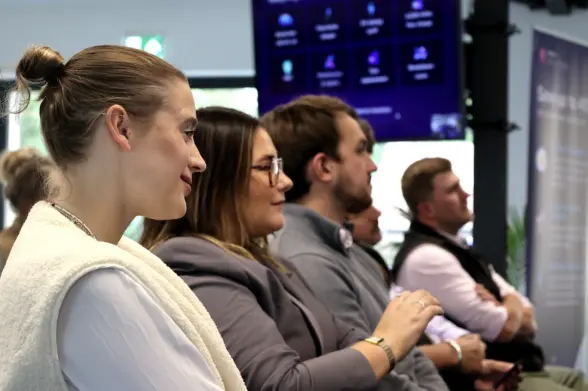 A group of professionals attentively listening during a presentation in a modern office setting. The focus is on three individuals in the foreground, with a screen displaying information in the background.