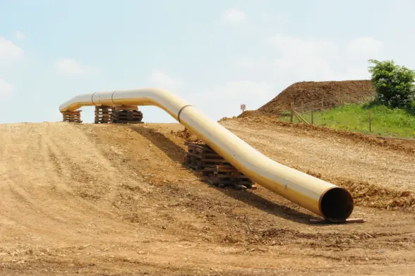 A large, curved pipeline resting on wooden pallets, running through a cleared, earthy landscape. The surroundings include mounds of soil and patches of grass under a clear sky.