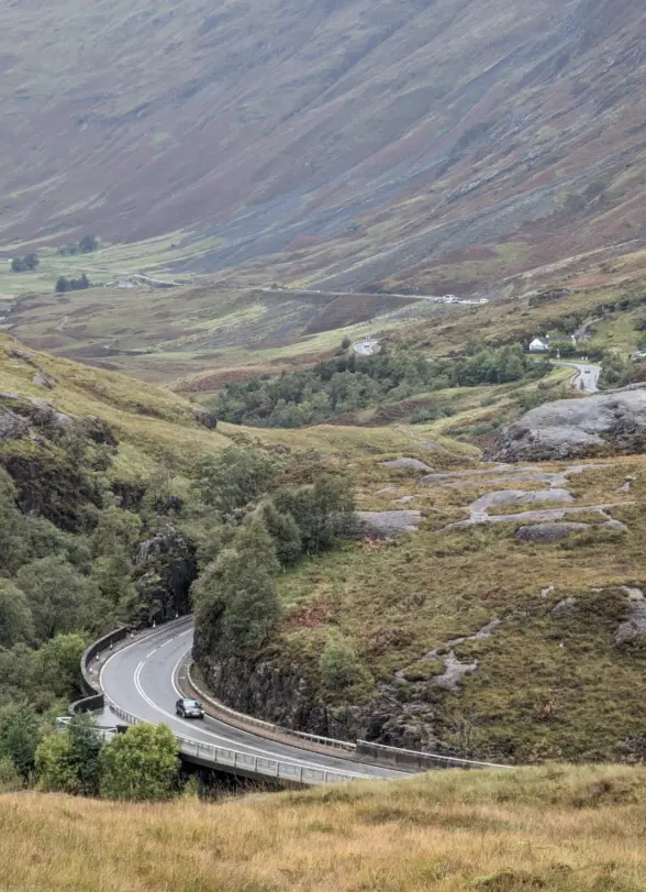 A winding road through a mountainous landscape, surrounded by green hills and rocky terrain. A few cars are visible on the road, with patches of grass and trees scattered throughout the valley.