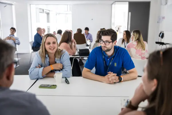 A group of people engaged in conversation in a modern office space. Two individuals at the foreground, one smiling and another looking attentively, sit at a large table with coffee cups and smartphones.