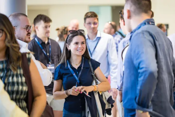 A group of people engaged in conversation at a conference or networking event. A young woman in a navy shirt and glasses is smiling while holding a phone. Others around her display varying expressions and attire.
