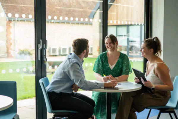 Three professionals engaged in discussion around a table in a bright office space, with large windows showing a grassy outdoor area. One person is writing notes, while another holds a tablet.