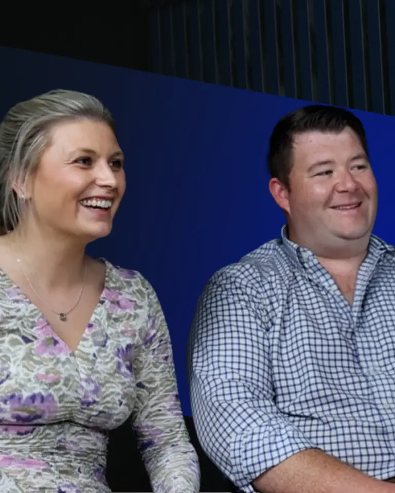 A woman in a floral dress and a man in a checkered shirt are seated, both smiling. They are in front of a blue background and appear to be engaged in a conversation.