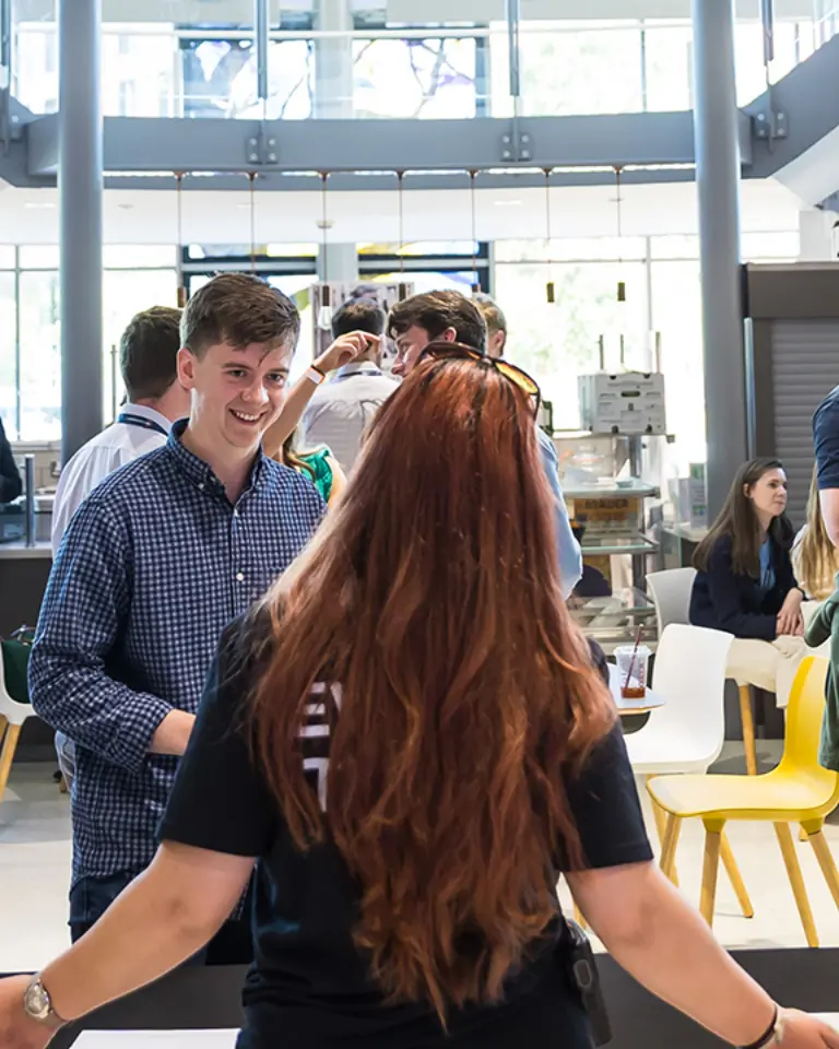 A group of young professionals networking in a modern atrium, with a woman in a black shirt welcoming attendees at a registration table. Bright windows and a staircase are visible in the background.