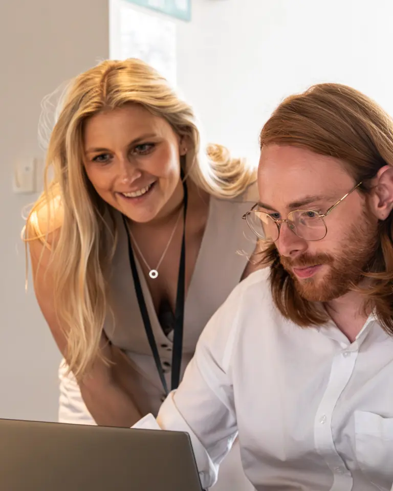 A woman standing beside a man seated at a laptop, both engaged in a discussion. The woman has long blonde hair and is wearing a light-colored dress. The man has long reddish-brown hair and glasses, focused on the screen.