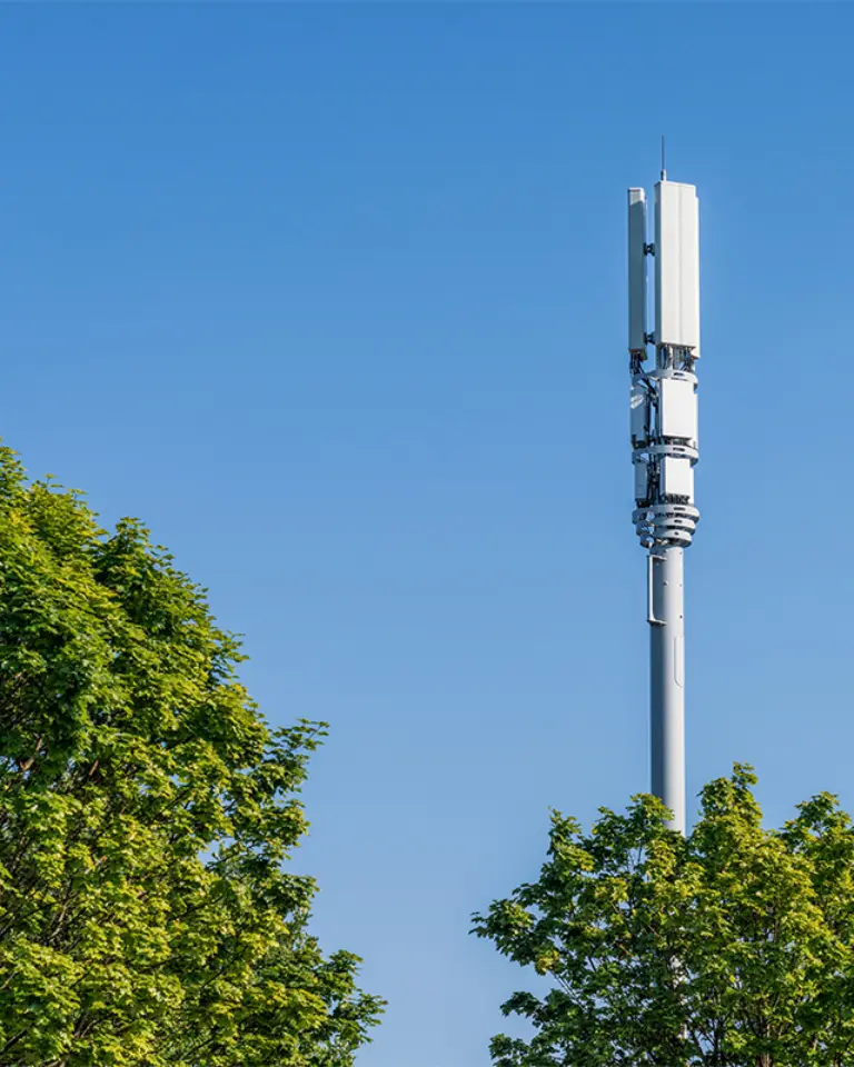 A telecommunications tower with antennas, surrounded by green trees against a clear blue sky.