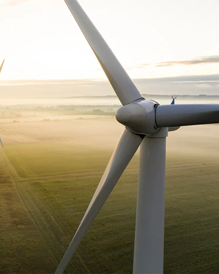 Two wind turbines standing in a green field during early morning, with a soft mist covering the ground and a light gradient in the sky.