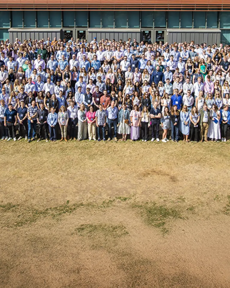 A large group of people, representing diverse demographics, gathered outside a modern building for a photo on a grassy area. They are dressed in casual attire and are arranged in multiple rows.